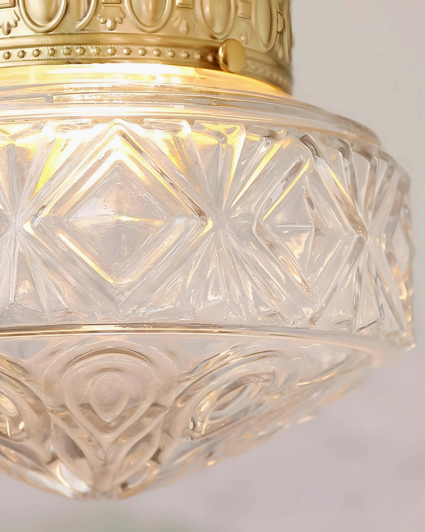 Close-up macro view of geometric carved clear glass ceiling lamp shade with brass gold metal trim, illuminated interior detail