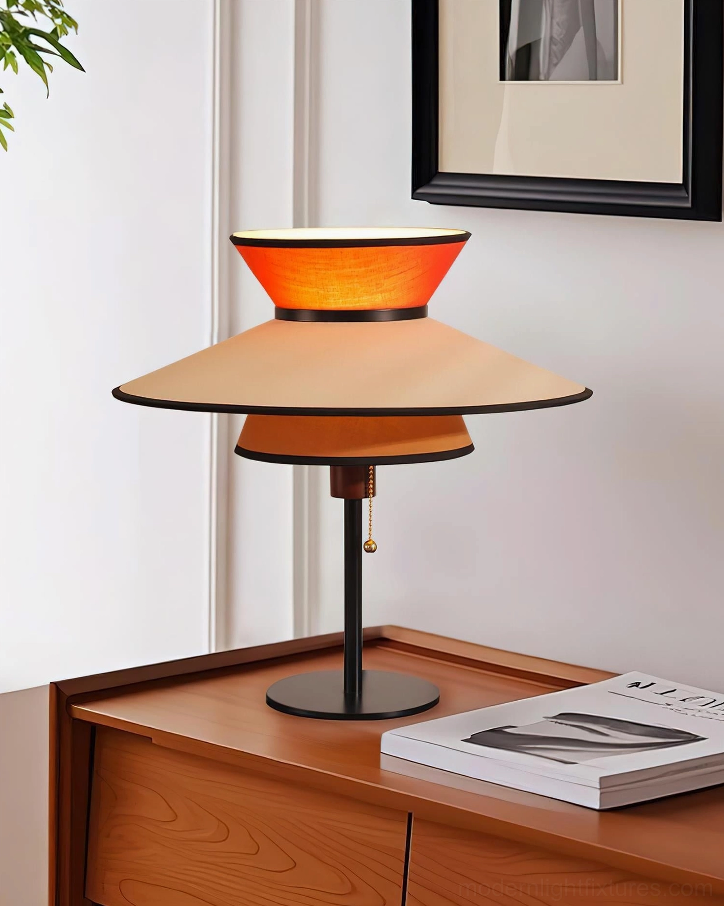 Modern geometric table lamp with three-tiered conical fabric shades on bedroom wooden dresser with stacked magazines and wall art
