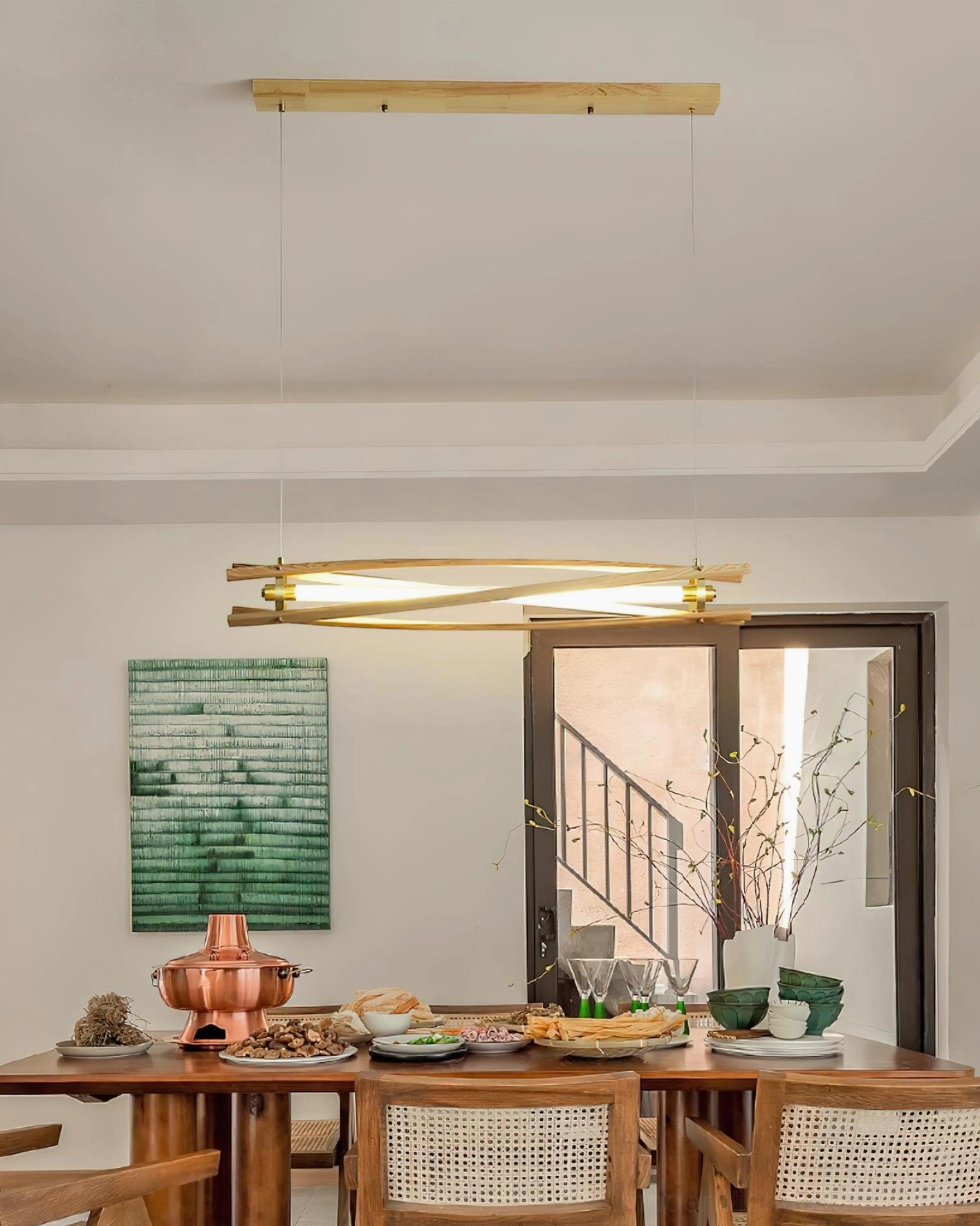 Natural wood linear chandelier above dining table with place settings and stairway backdrop in contemporary home