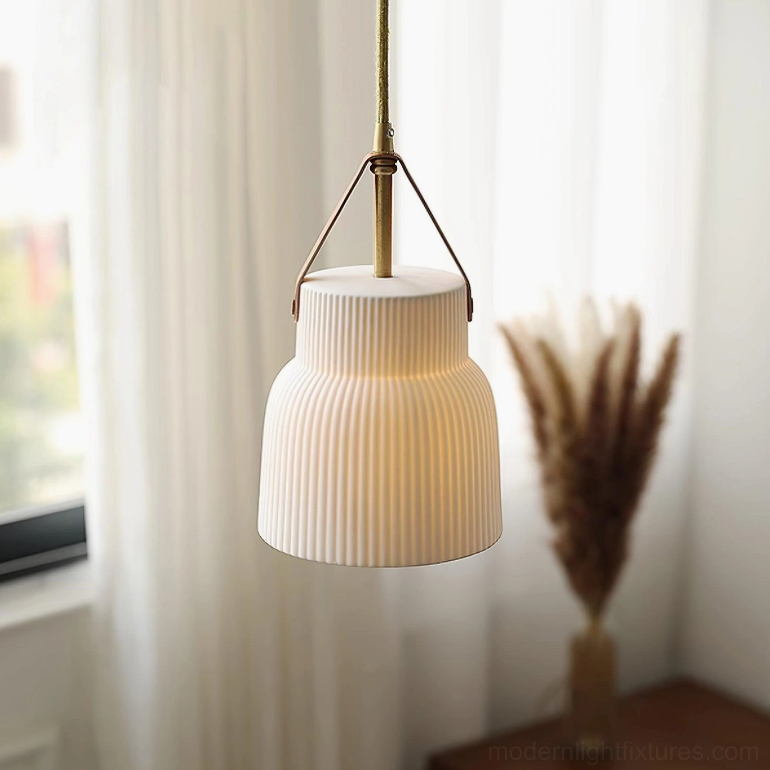 Three-quarter view of tiered ceramic ridged pendant with brown leather strap and brass hardware against windowsill with pampas grass