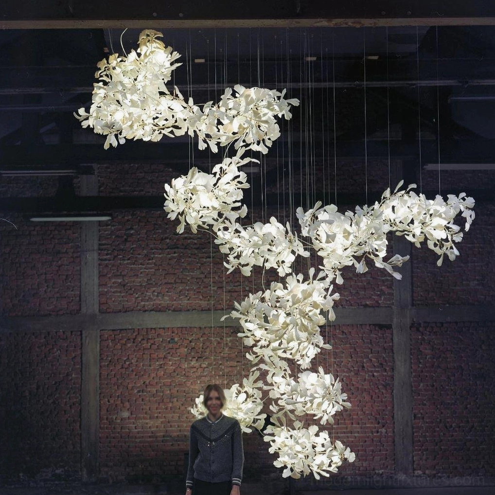 Large-scale ginkgo leaf chandelier in brick industrial hall with human figure showing scale and warm glowing light