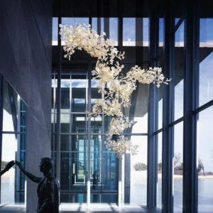 Modern ginkgo chandelier installed in contemporary glass atrium with warm glowing light illuminating organic leaf structure