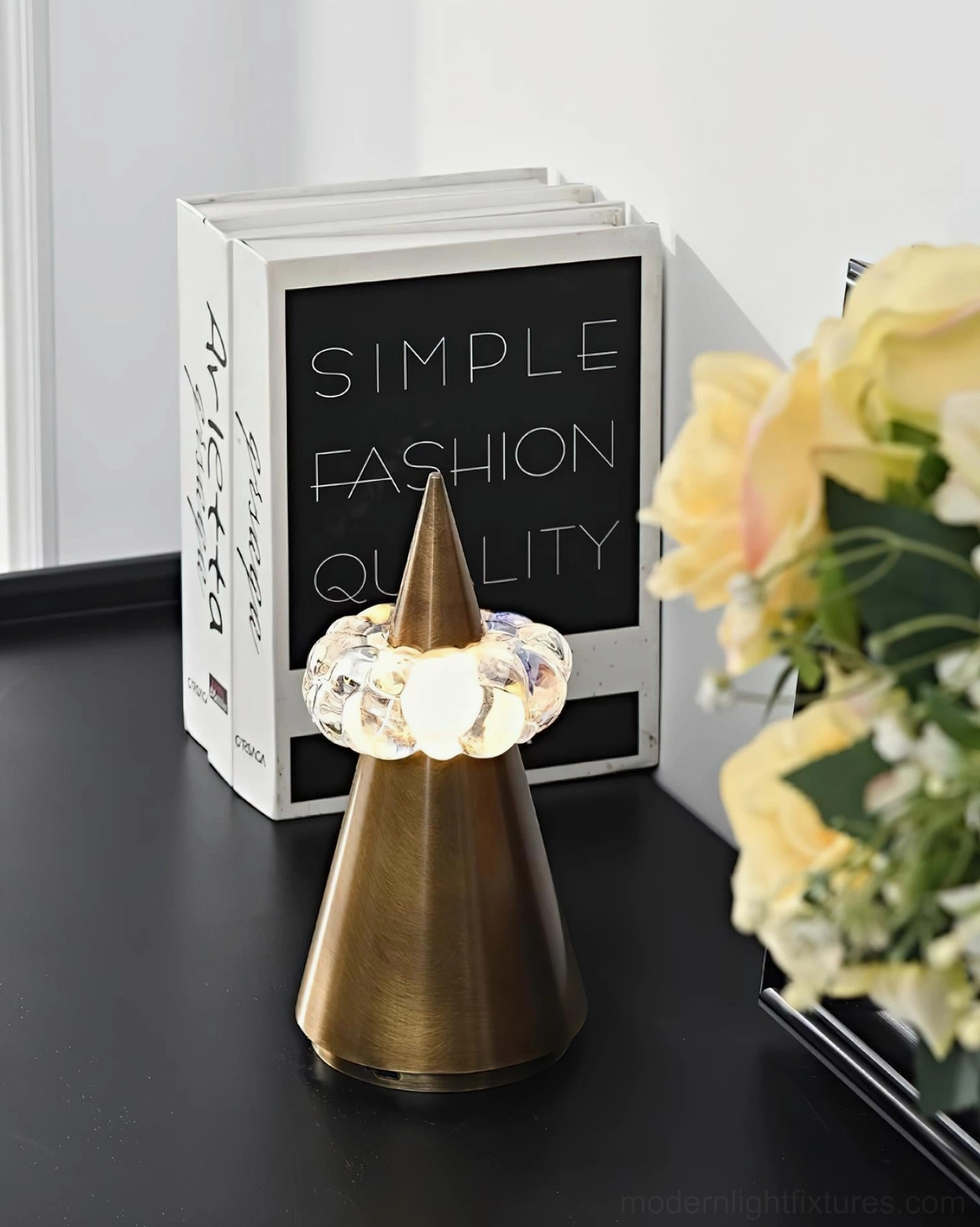 Brushed bronze sculptural table lamp on black side table with decorative books and fresh floral arrangement, warm ambient lighting