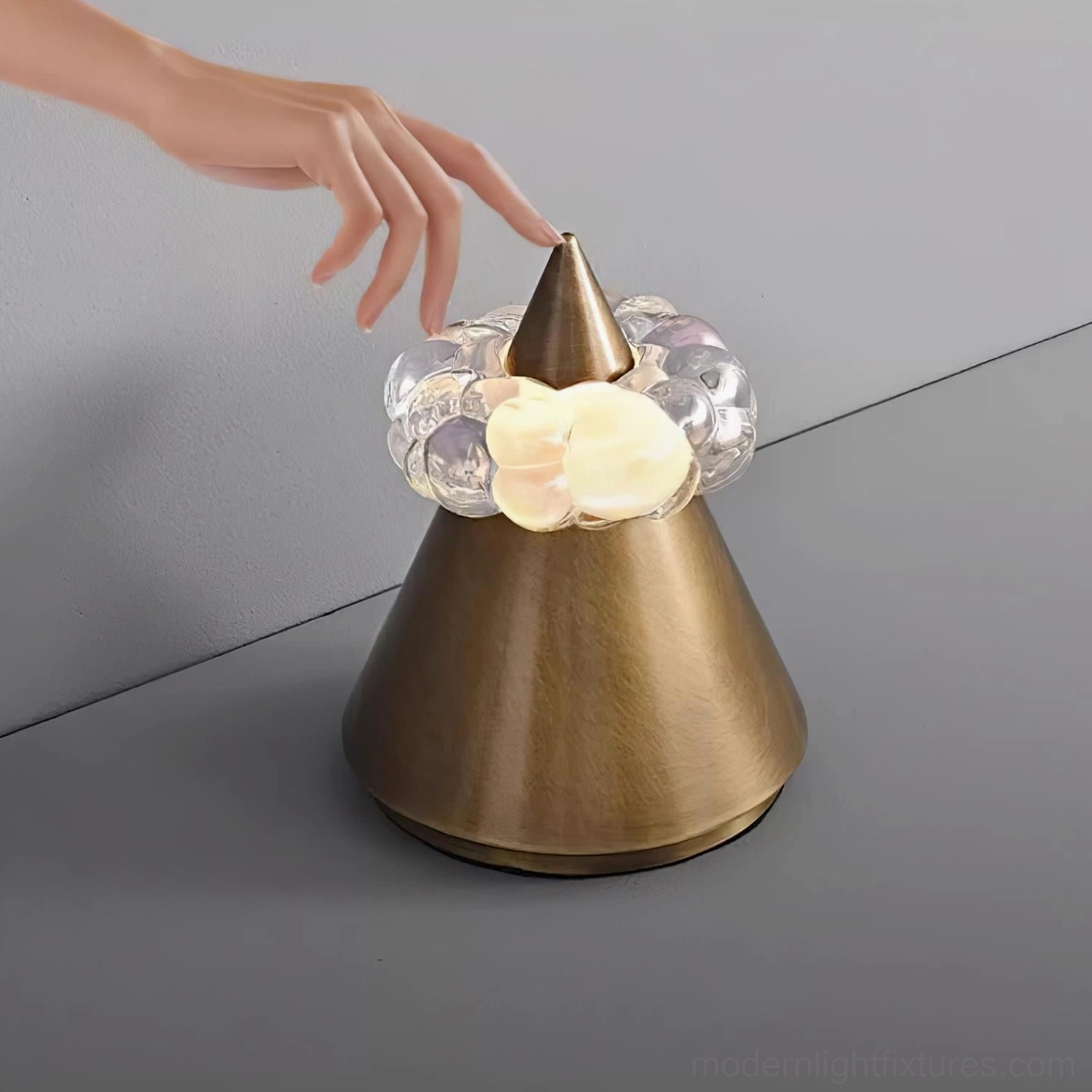 Close-up detail of Glint table lamp cone and crystal light ring, hand touching metal body, soft golden ambient glow on gray studio backdrop