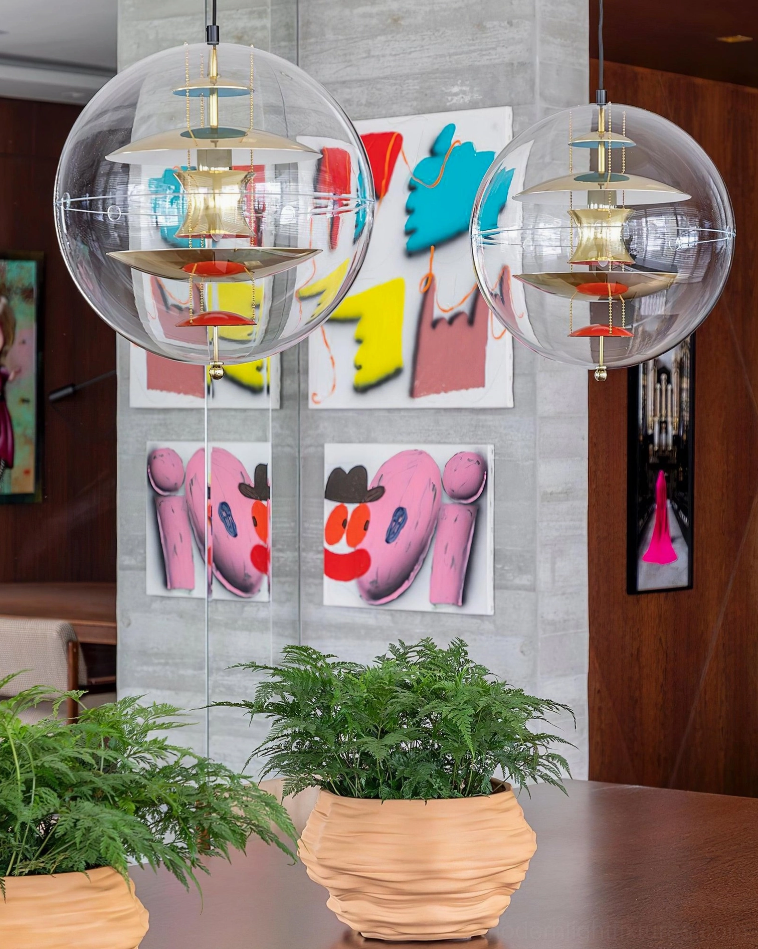 Two modern globe pendant lights suspended above wooden dining table in contemporary dining room with potted plants and wall art