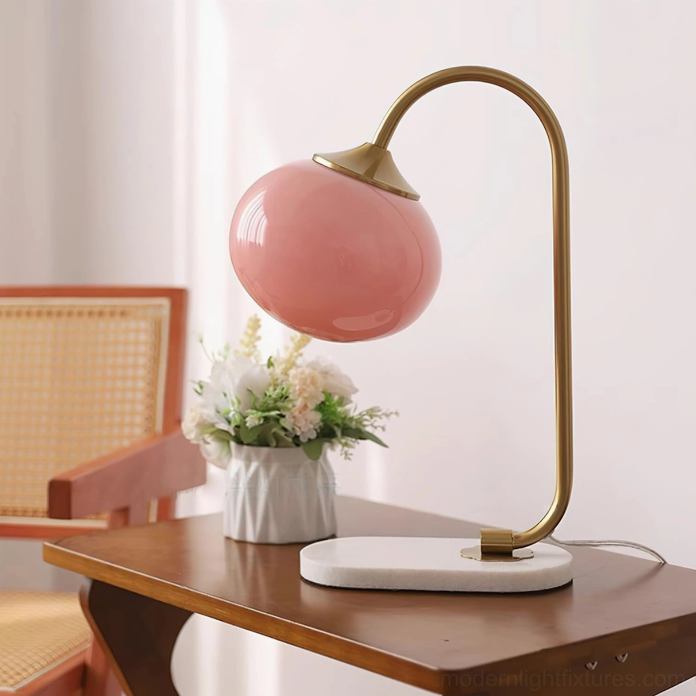 Globe table lamp with pink glass shade and gold brass curved arm displayed on side table with floral vase decor