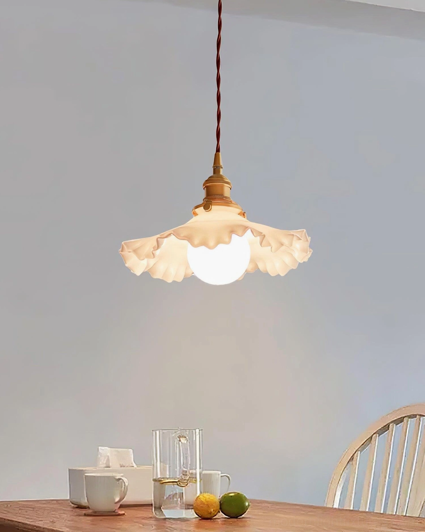 Ruffled glass pendant light with glowing bulb suspended over wooden dining table with tableware and twisted fabric cord