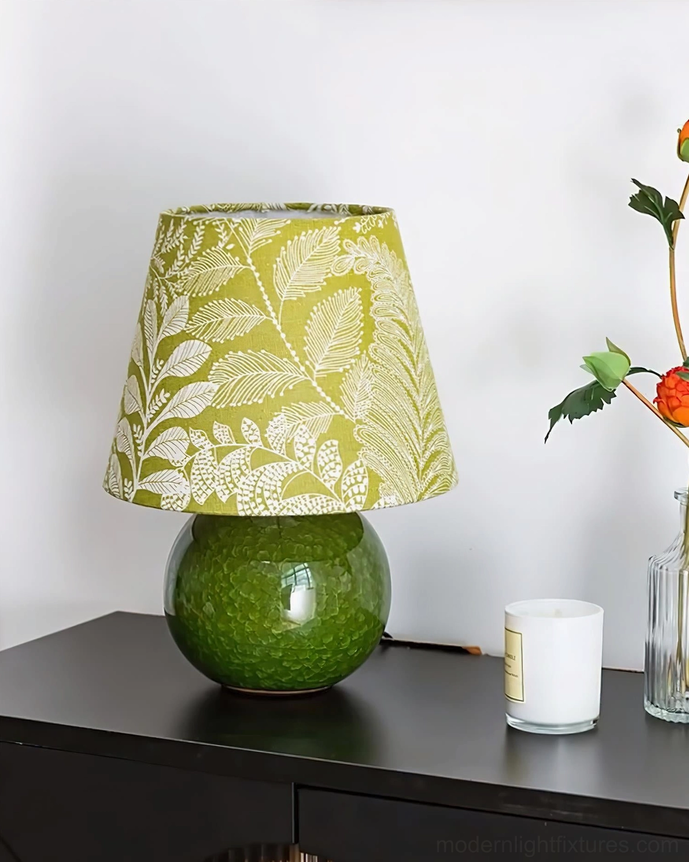 Green foliage-print table lamp on dark cabinet paired with floral vase and candle