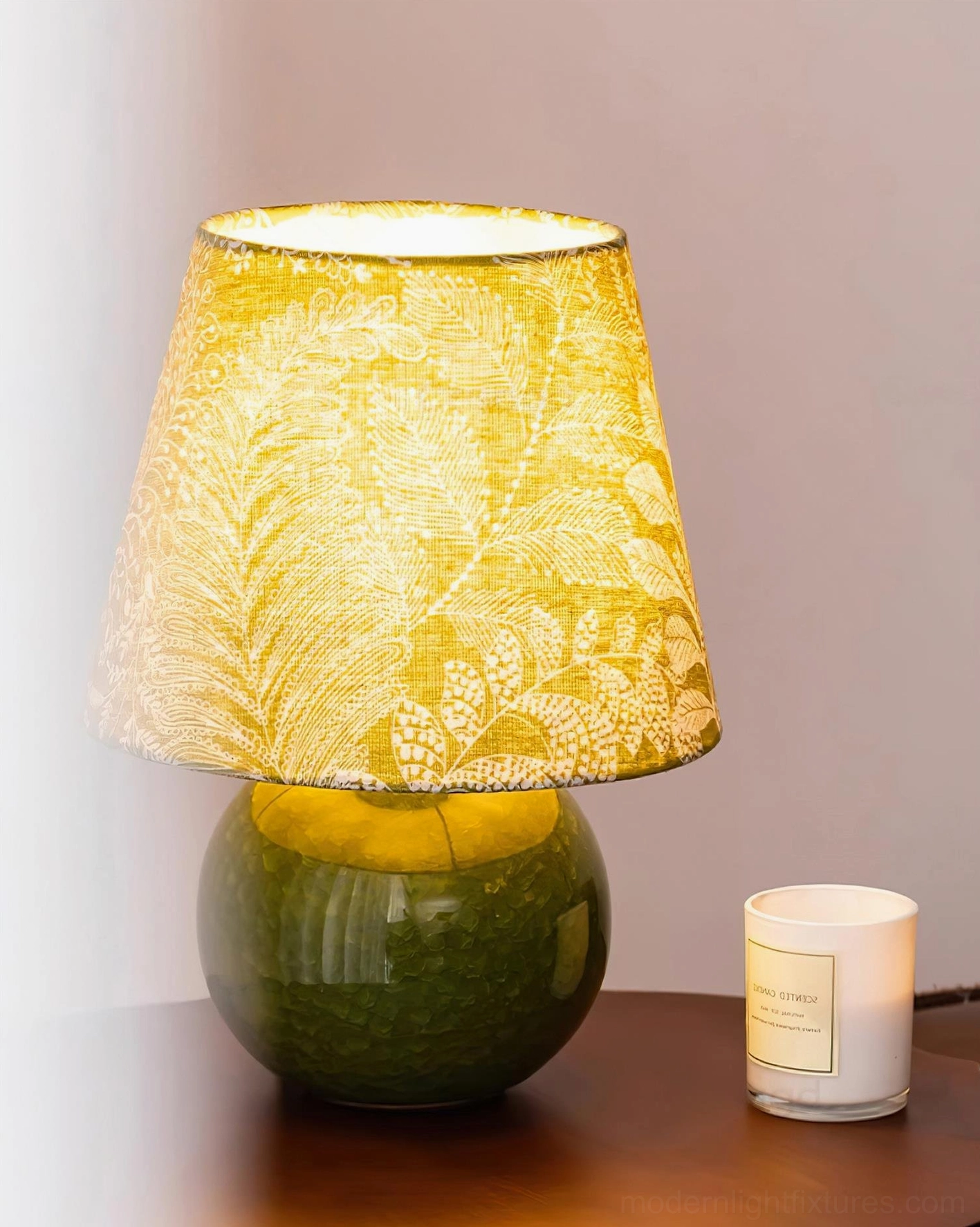 Green botanical table lamp glowing beside candle on wooden side table