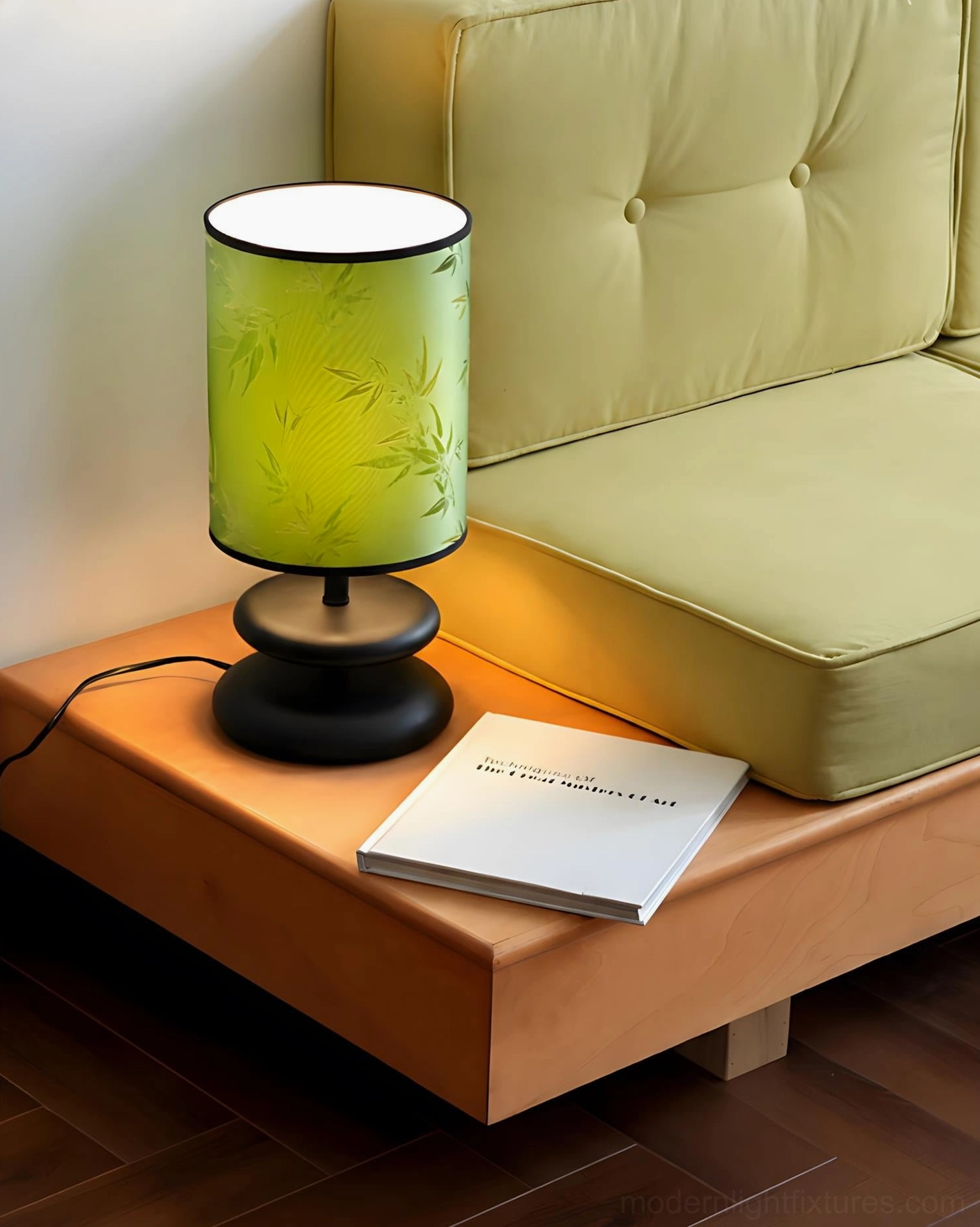 Green cylindrical table lamp with bamboo leaf print, dark wood and gold base, side three-quarter view on sofa bench