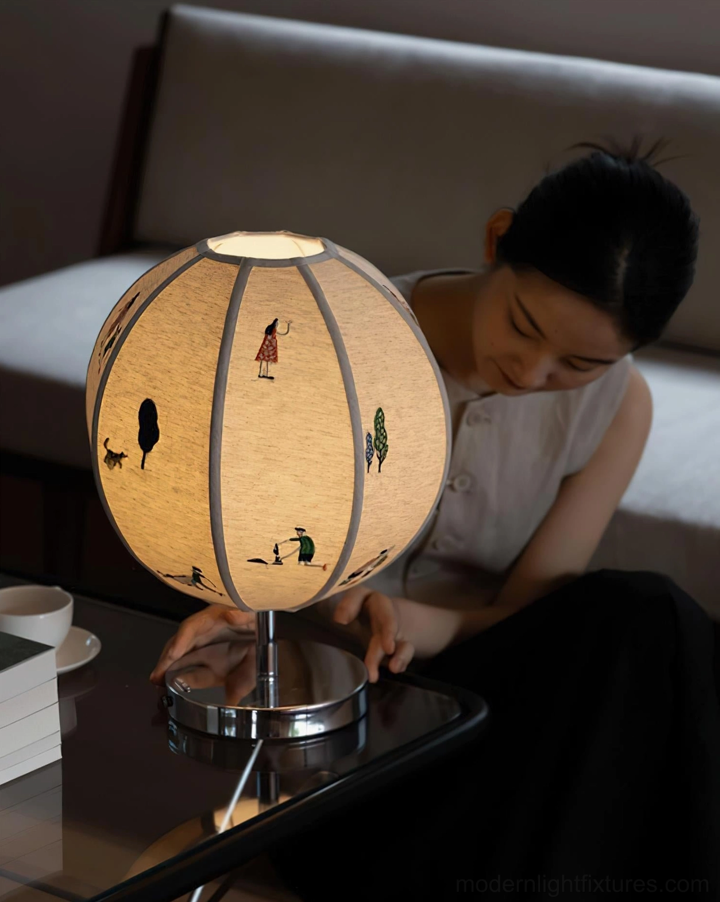 Woman adjusting illuminated embroidered narrative table lamp in living room, front three-quarter view showing warm glow and cartoon motif design