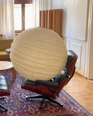 Off-white ribbed paper pendant lamp suspended above Eames lounge chair in mid-century modern living room with warm neutral decor