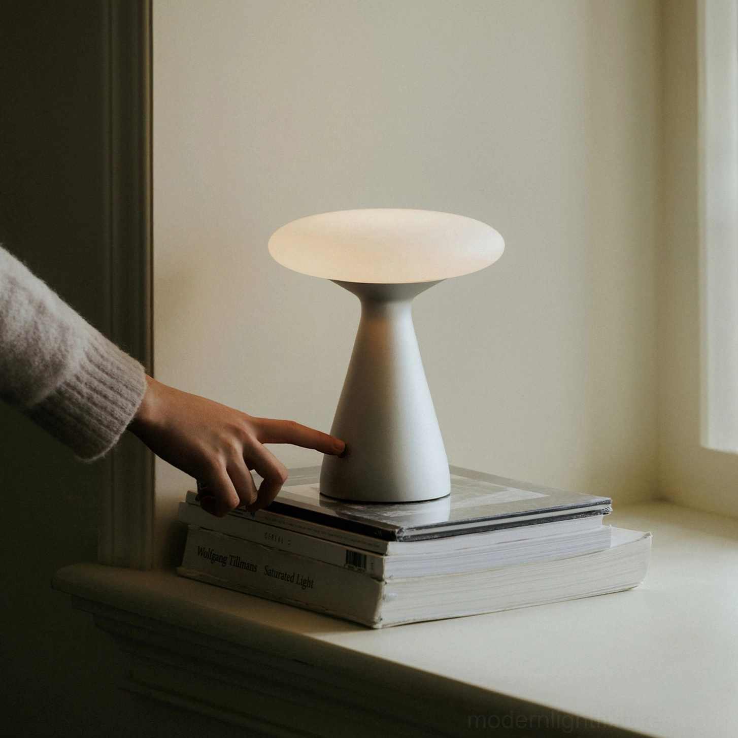 Konos cordless table lamp on residential windowsill with stacked books - hand touching base to activate touch switch for color changing light