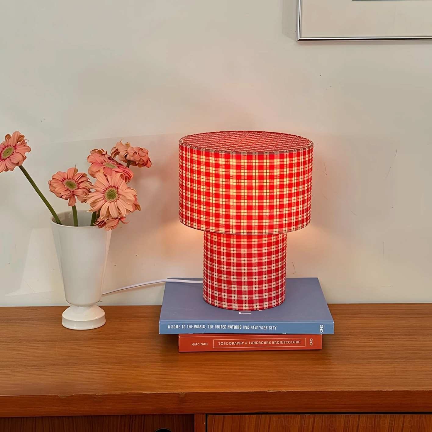 Lit red gingham latticefield table lamp on wooden side table with stacked books and flower vase home interior