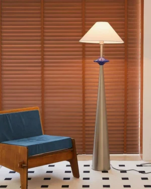 Lazuro modern floor lamp displayed in contemporary living room beside blue upholstered armchair, three-quarter view with wooden blinds