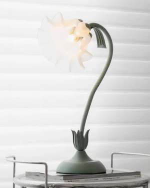 Modern lily flower table lamp illuminated on side table with louvered window backdrop showing curved green arm