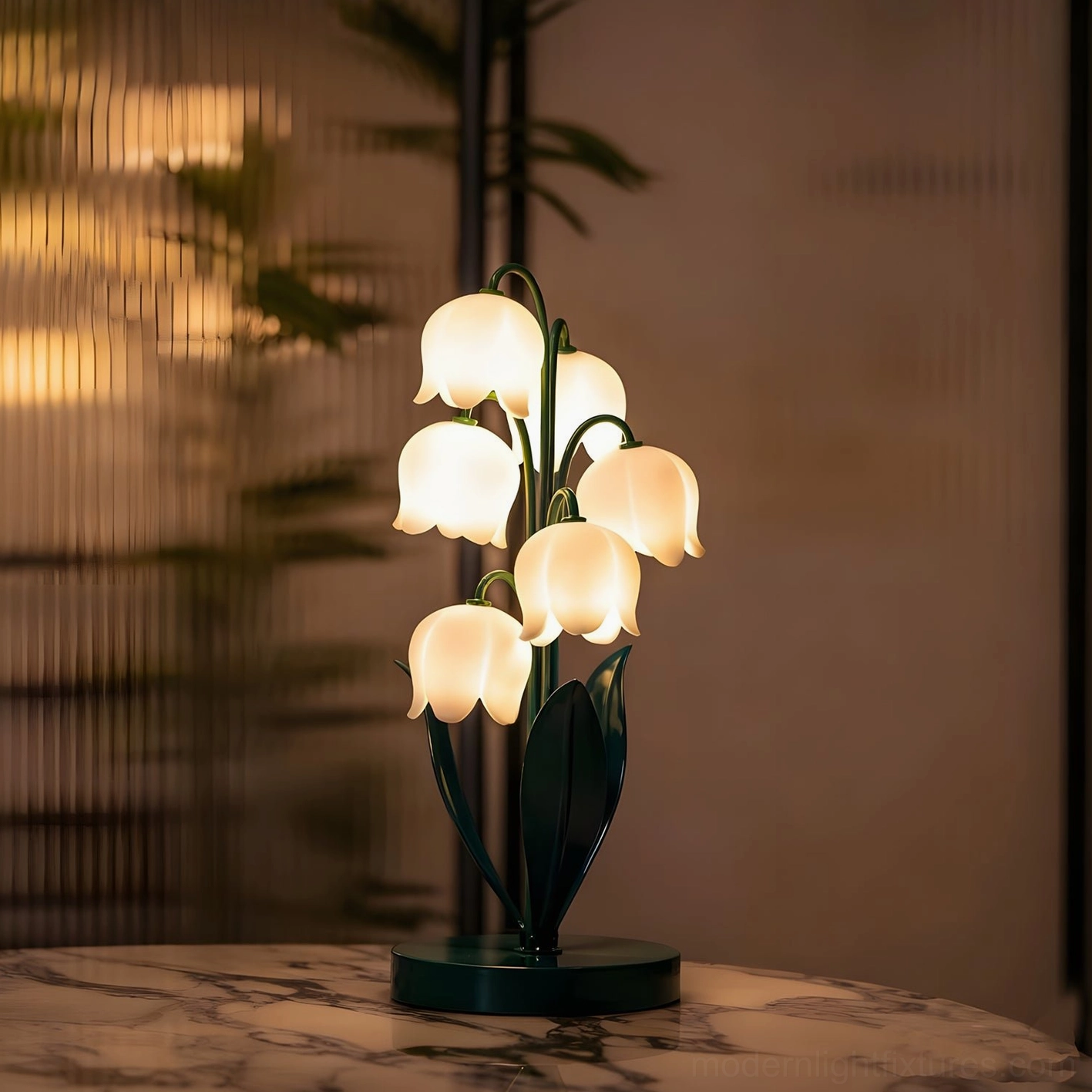 Modern botanical table lamp with dark green metal components on marble tabletop, warm glow with blurred foliage background creating cozy home scene