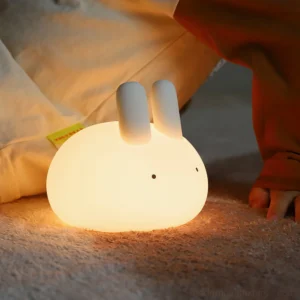 Lit bunny night light with warm white 3000K glow on bedroom carpet near bed linens