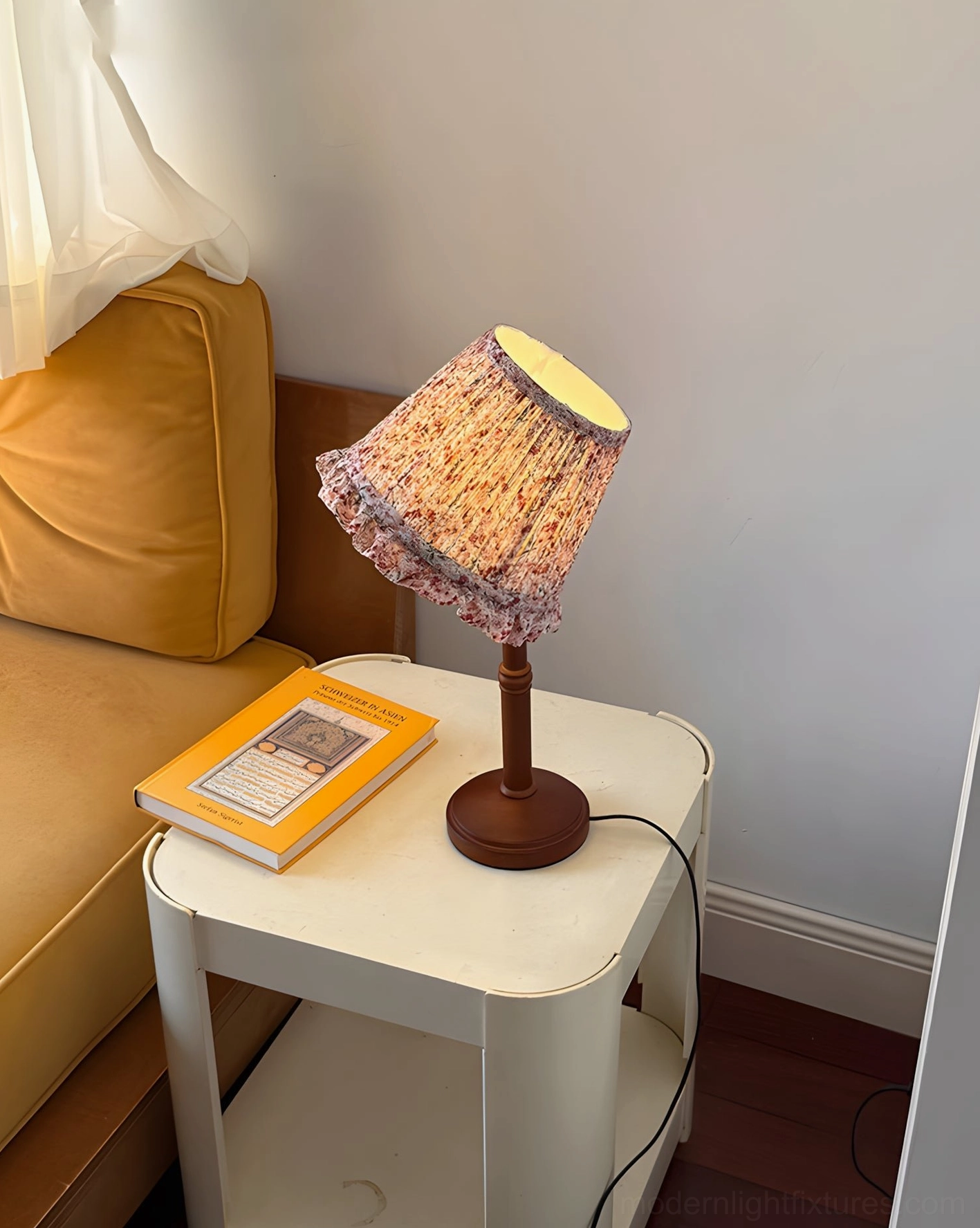 Lit cottage floral lamp on white side table beside tan fabric sofa with hardcover book, warm glow