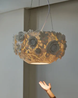 Illuminated ceramic flower pendant lamp hanging at eye level with warm glowing light, suspension wires visible against minimal wall