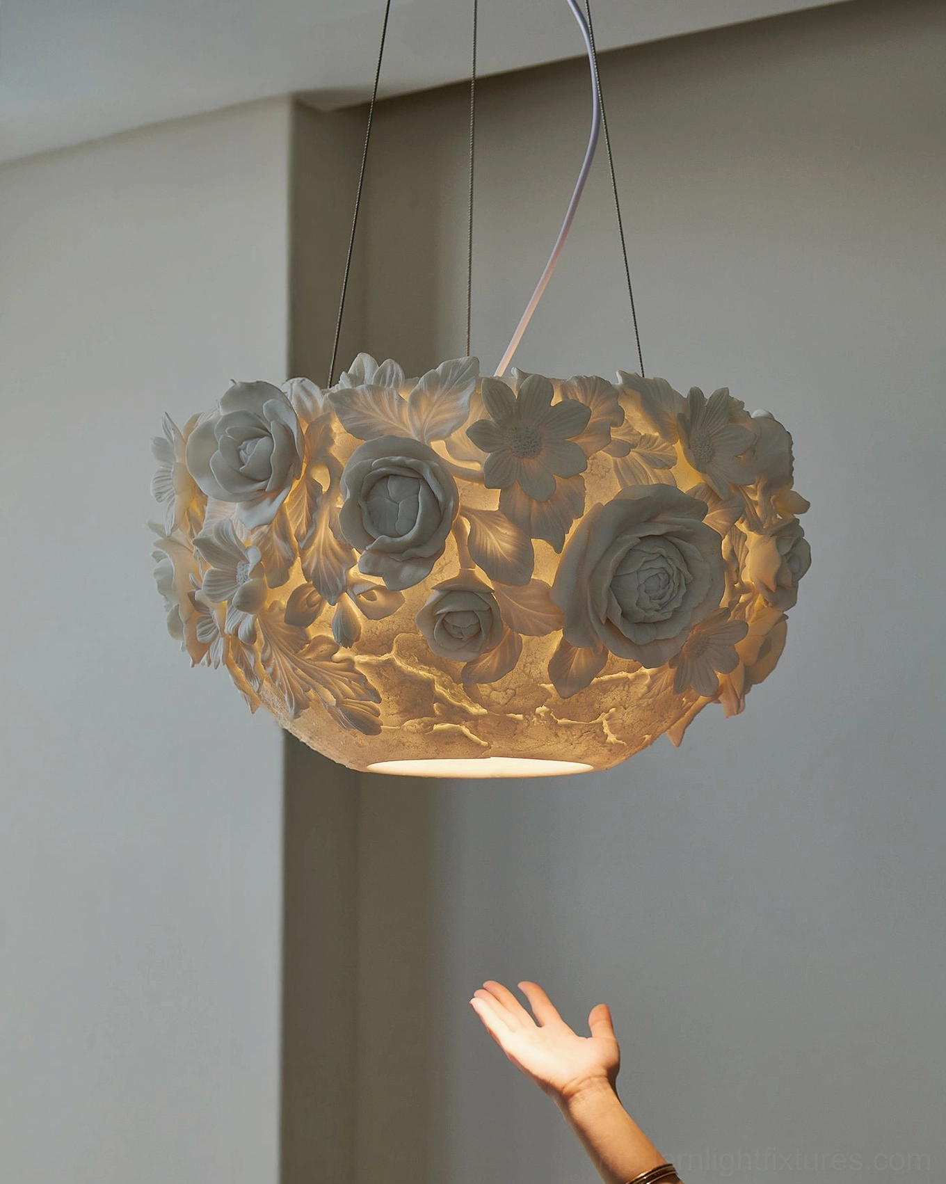 Illuminated ceramic flower pendant lamp hanging at eye level with warm glowing light, suspension wires visible against minimal wall