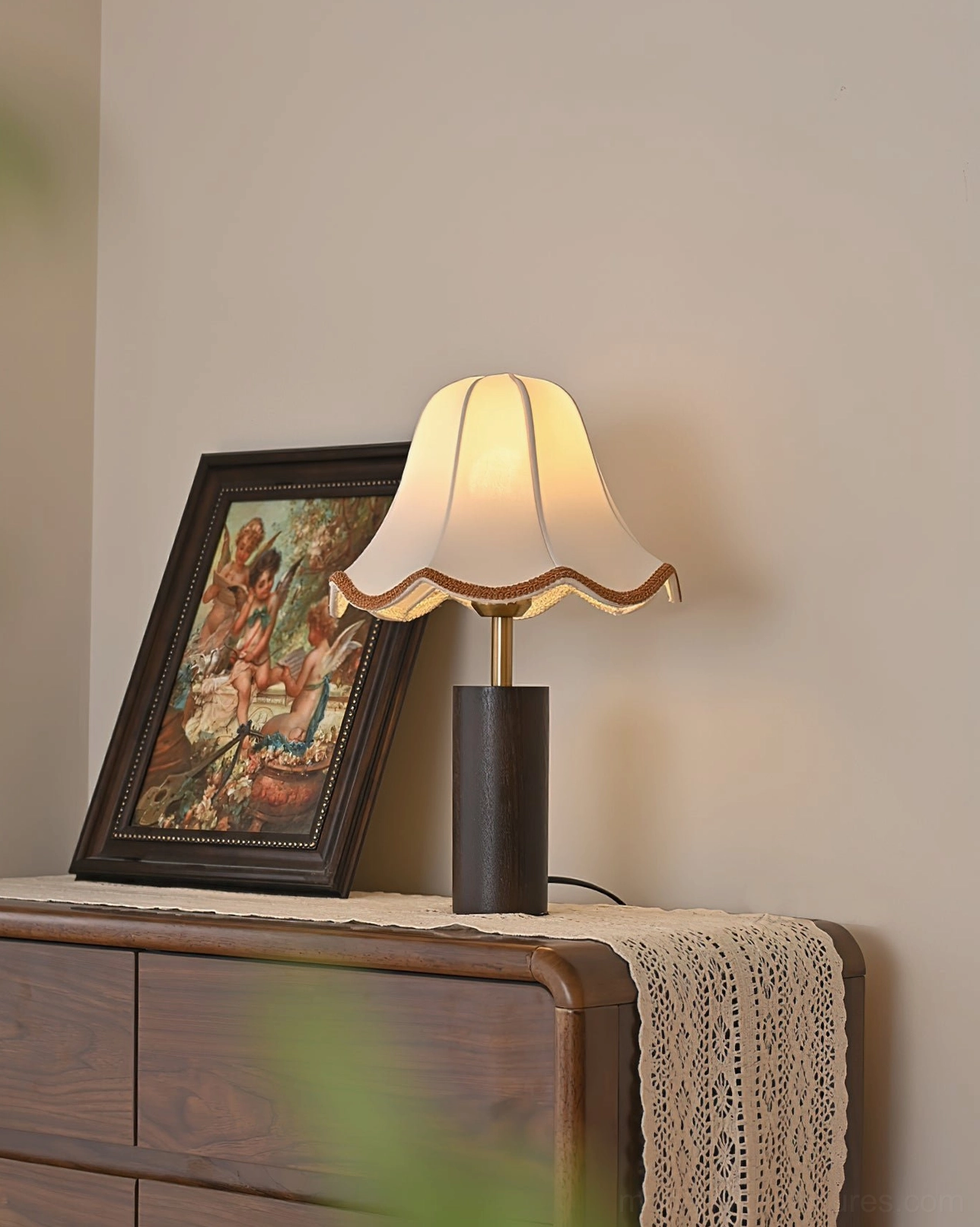 Lit table lamp glowing on wooden dresser with lace runner and vintage framed art three-quarter view