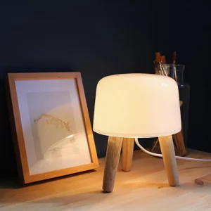 Illuminated modern milk glass table lamp with warm glow in cozy home corner setting, paired with wooden floor and framed artwork on wall