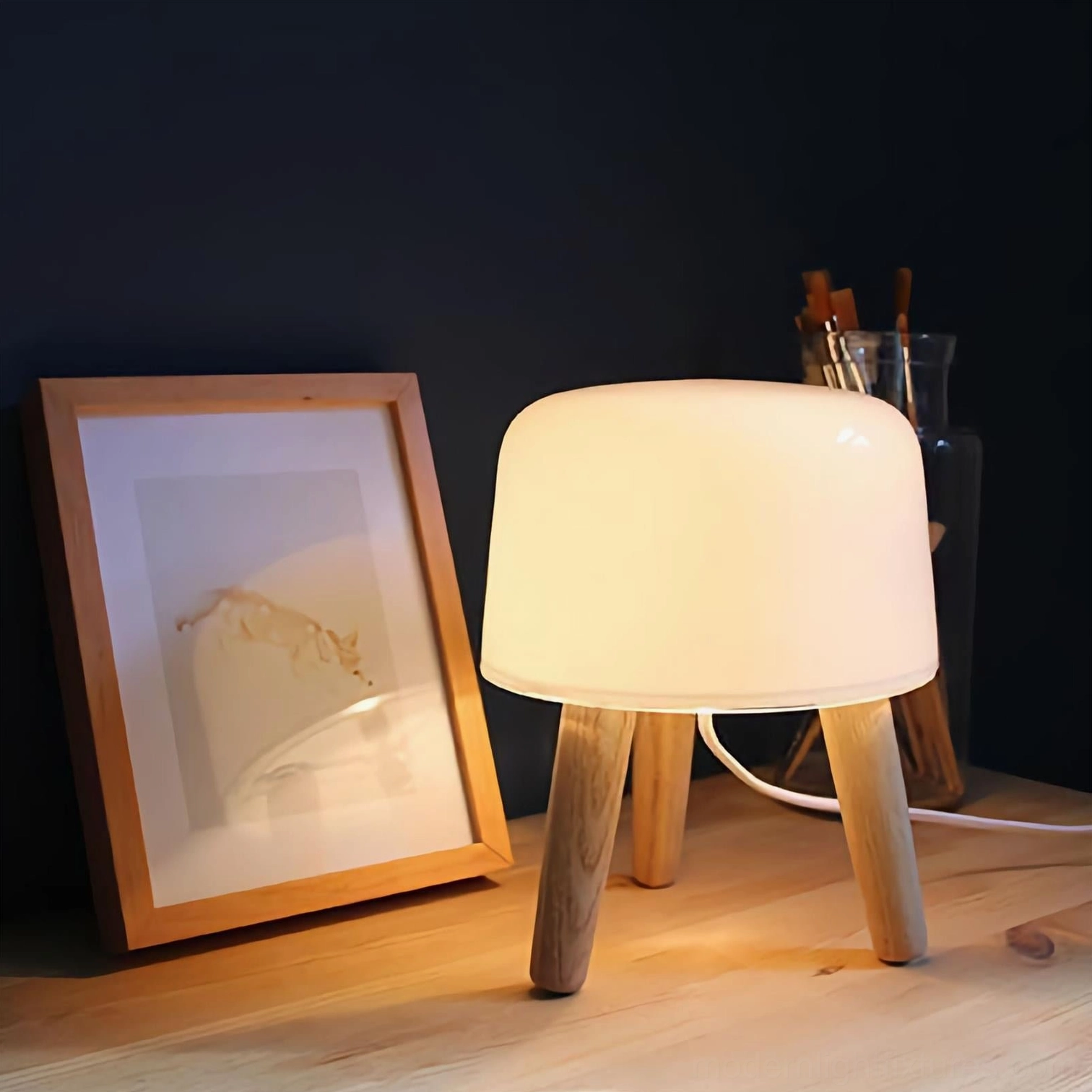 Illuminated modern milk glass table lamp with warm glow in cozy home corner setting, paired with wooden floor and framed artwork on wall