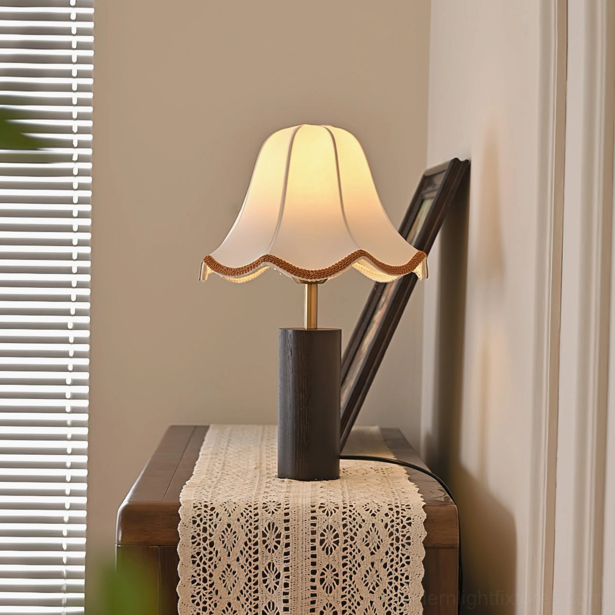 Lit table lamp with glowing scalloped shade on bedroom side table with lace runner and window blinds