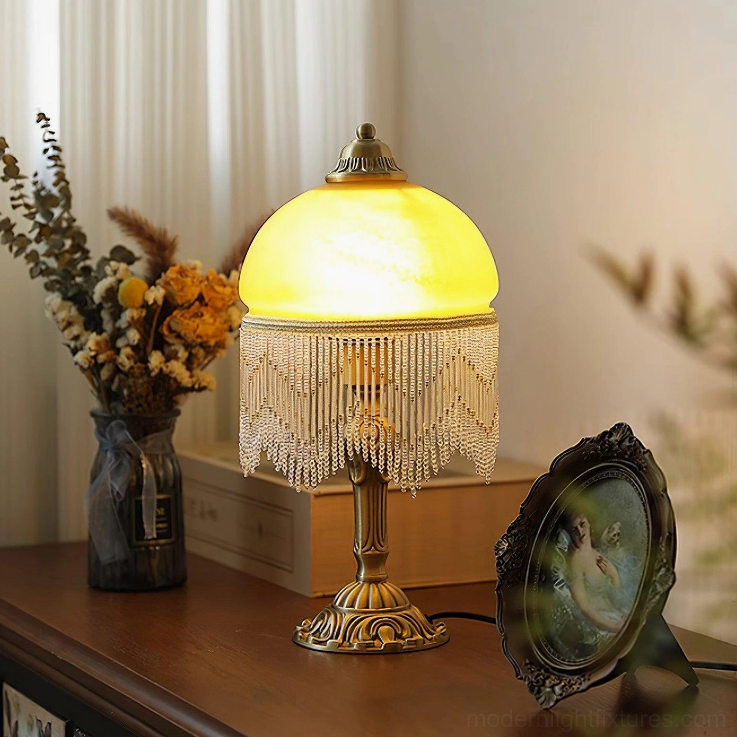 Lit vintage brass table lamp with warm amber frosted glass shade and crystal beaded tassel on wooden surface with dried flowers