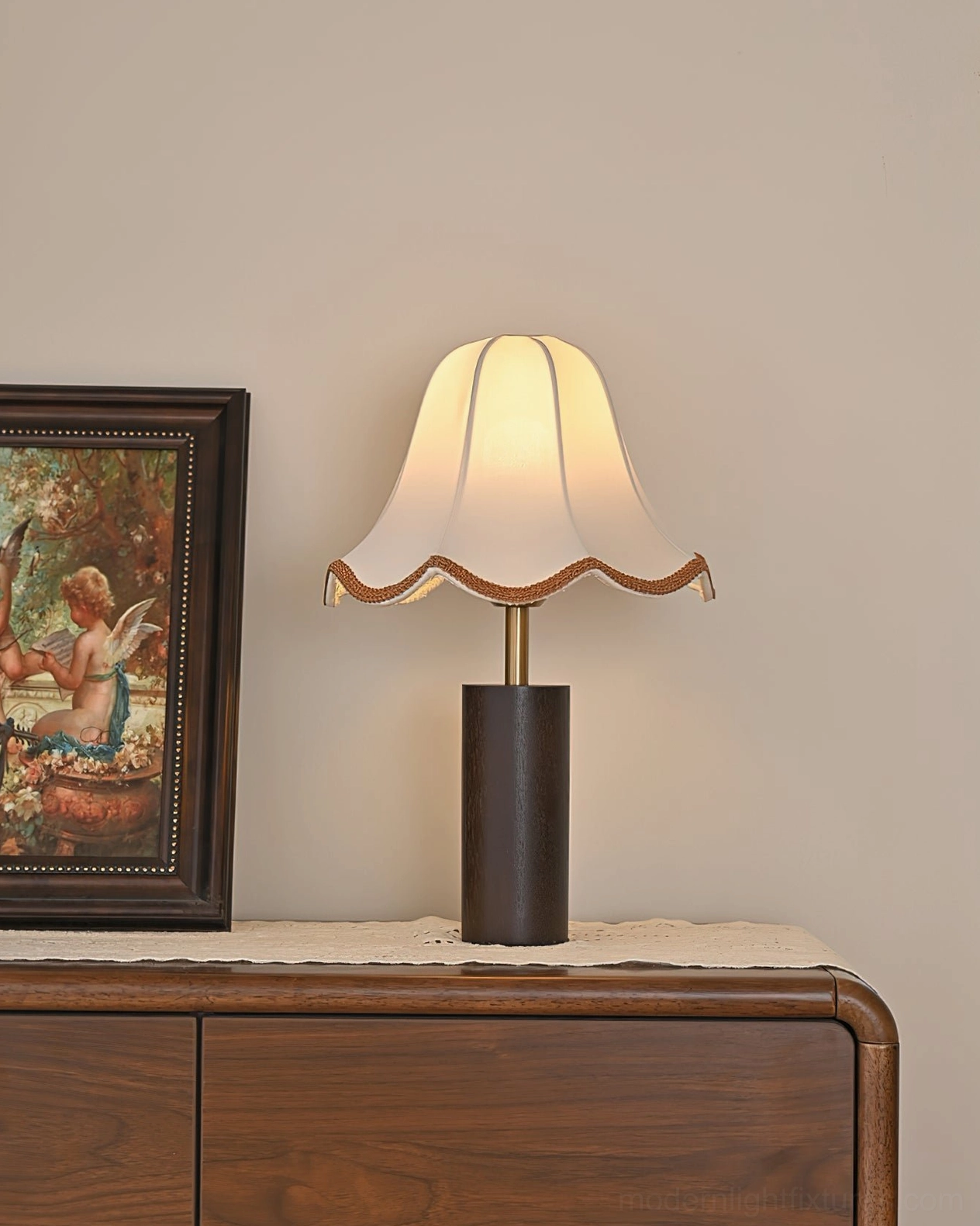 Lit walnut table lamp with scalloped fabric shade against wall interior with vintage framed art