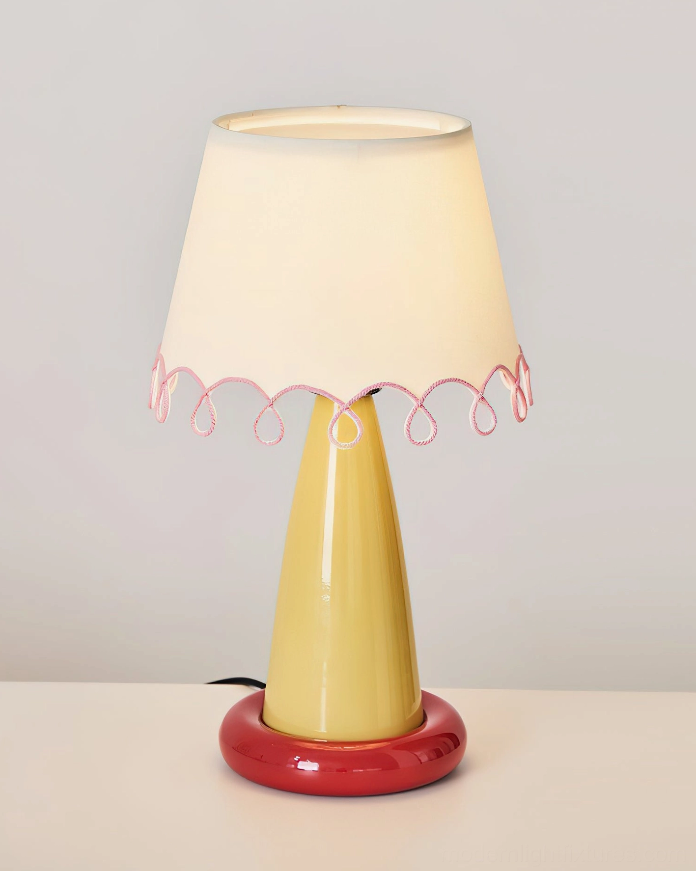 Illuminated Lollio ceramic table lamp featuring yellow stem, red base, and glowing white fabric shade with pink trim accent