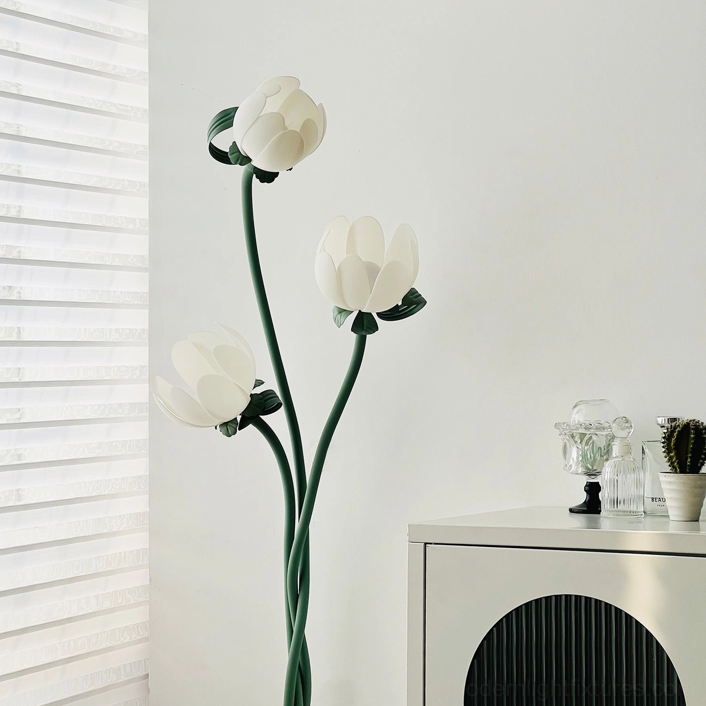 Three-head magnolia floor lamp with curved green stem and white acrylic flower shades in sunlit room with louvered window