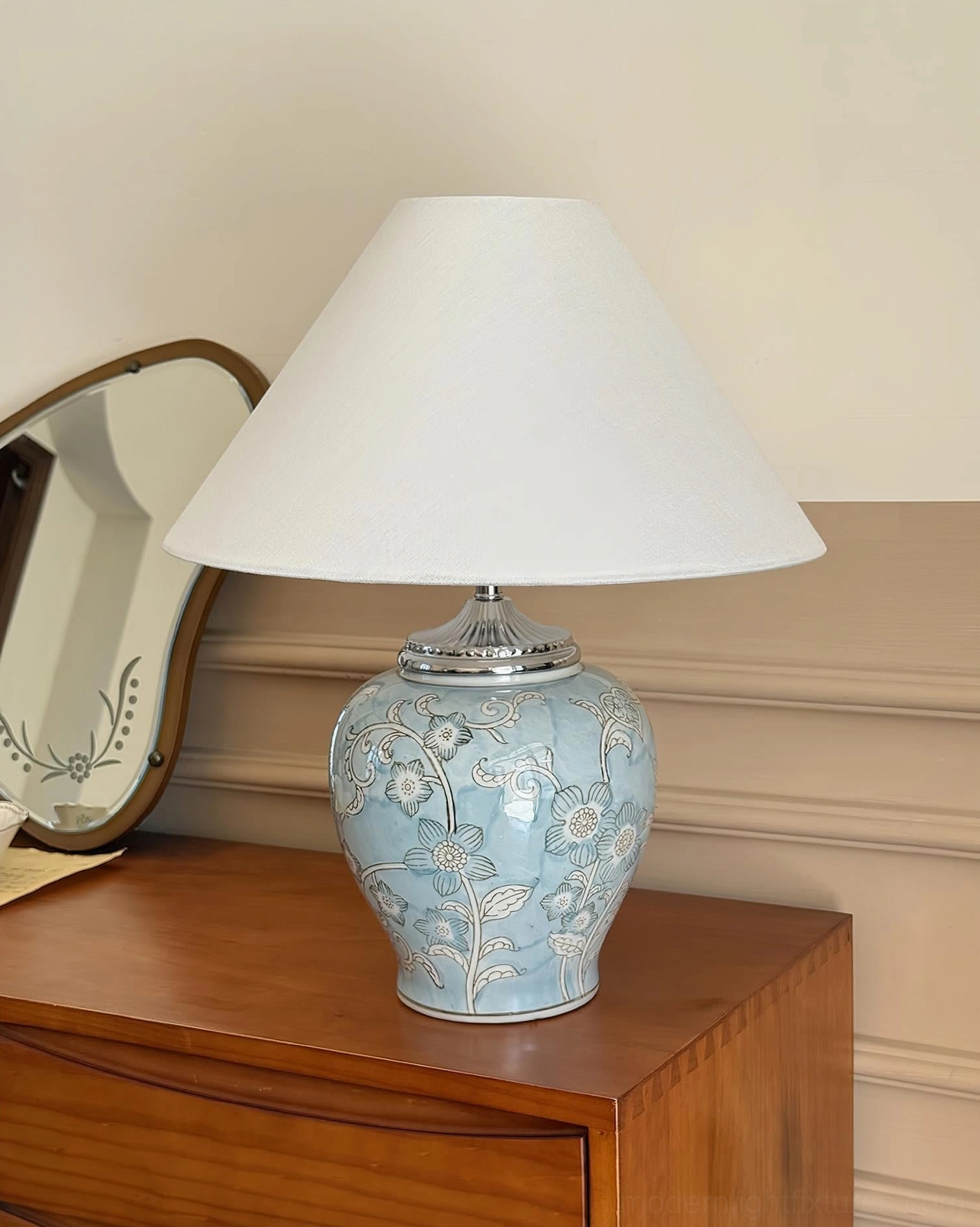 Light blue hand-glazed ceramic table lamp with white floral prints and fabric shade displayed on bedroom dresser with vintage wall mirror and decor accessories