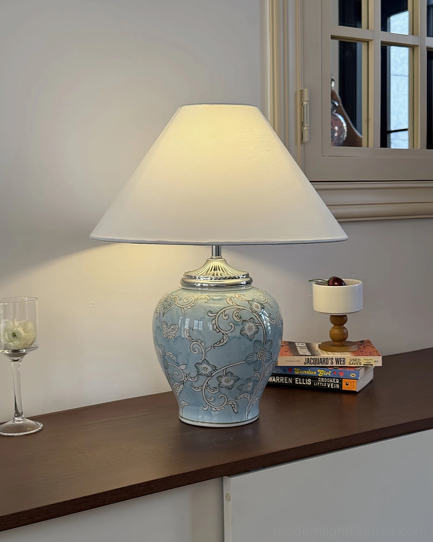 Pale blue ceramic table lamp with embossed florals and off-white fabric shade styled on living room console table with wine glass and art books, contemporary interior arrangement