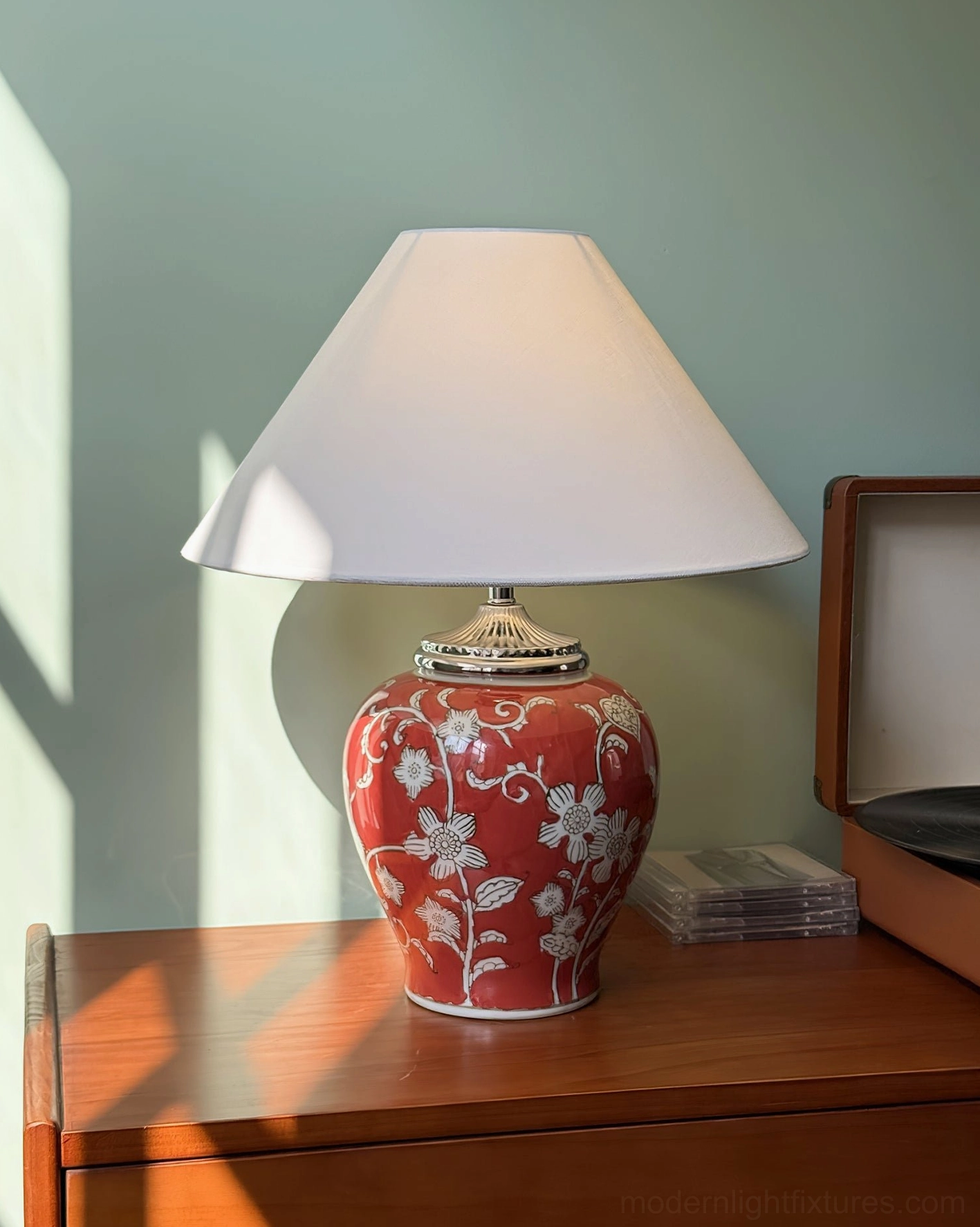 Crimson red hand-glazed ceramic table lamp with white floral medallion designs and white fabric shade on sunlit wooden side table with decorative accessories and natural lighting