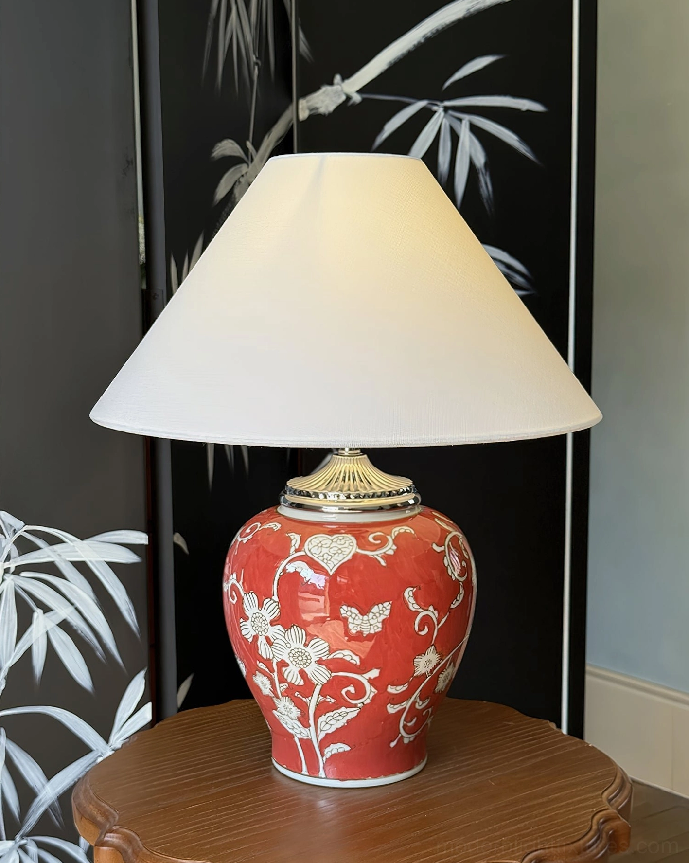 Bright red ceramic table lamp featuring white floral motifs on hand-glazed base with white fabric lampshade, displayed on round wooden side table against bamboo screen backdrop