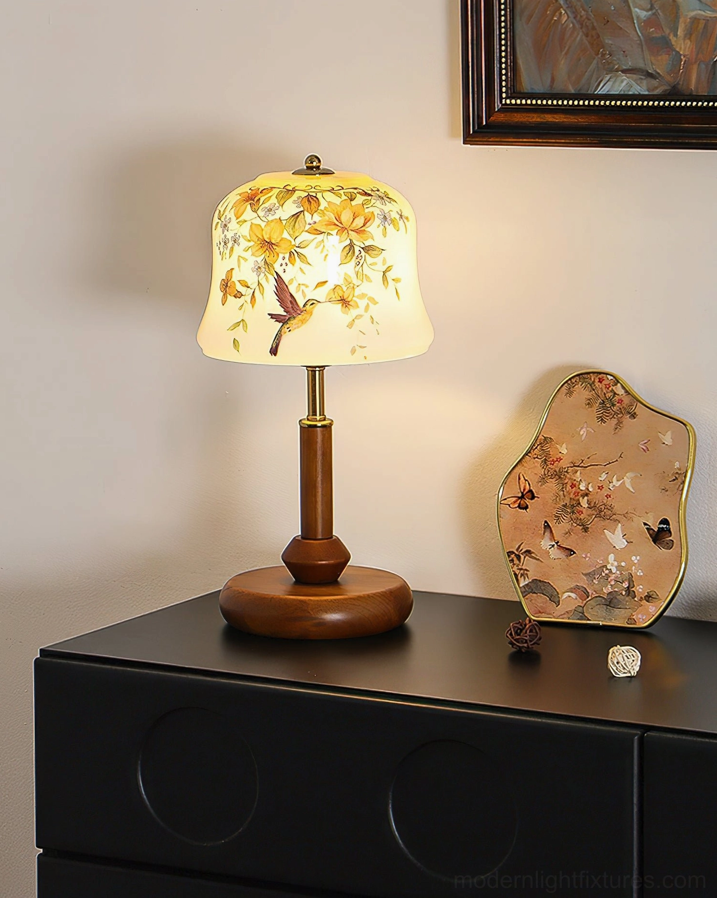 Illuminated Meadow table lamp with warm glowing light showing floral pattern, positioned on black dresser with home décor