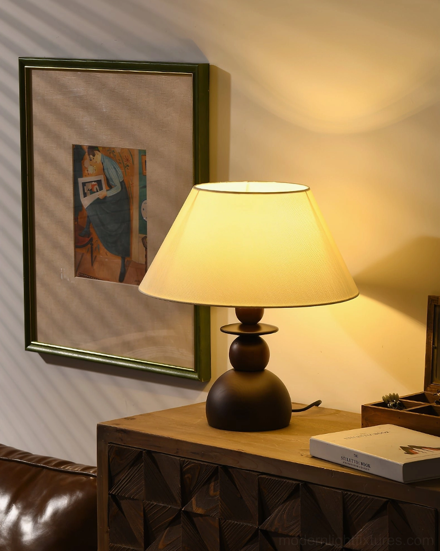 Illuminated fabric shade table lamp on dresser next to leather sofa with book decor