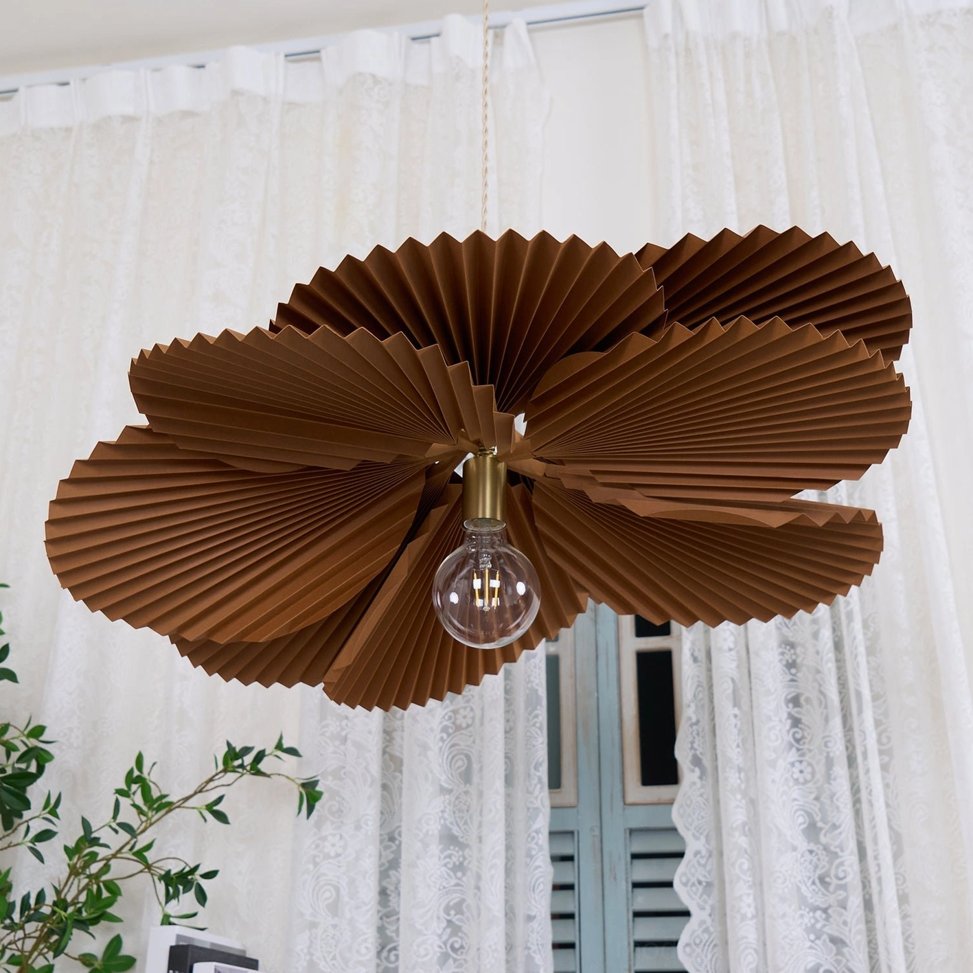 Brown pleated pendant fixture suspended in vintage interior with lace curtain and window showing warm ambient lighting