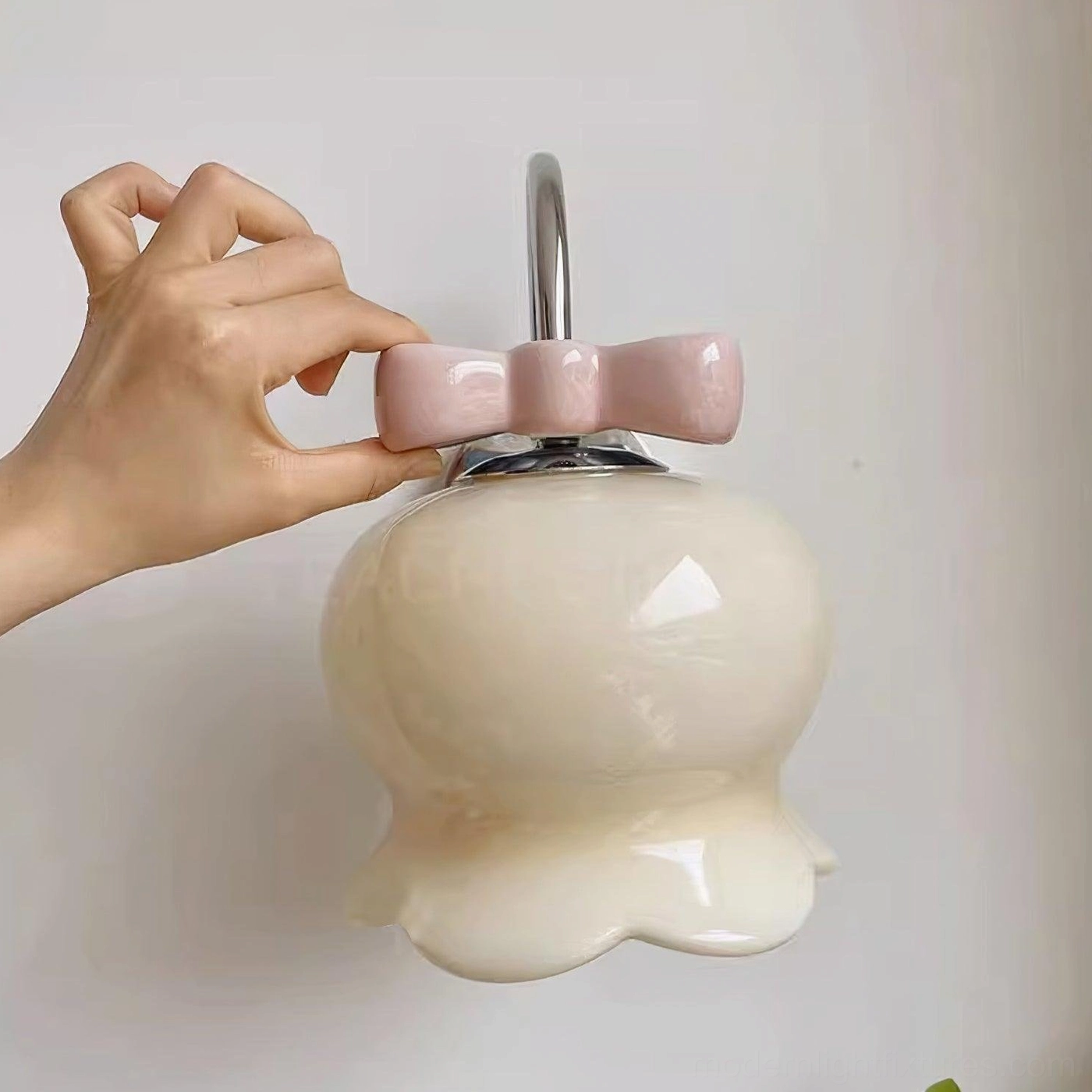 Close-up handheld detail view of Millie Bells wall sconce with cream ceramic shade, chrome metal top hardware, and pink bow accent