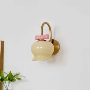 Cream yellow ceramic Millie Bells wall sconce with brass metal mounting bracket and pink bow detail, displayed in minimalist home interior with green plants