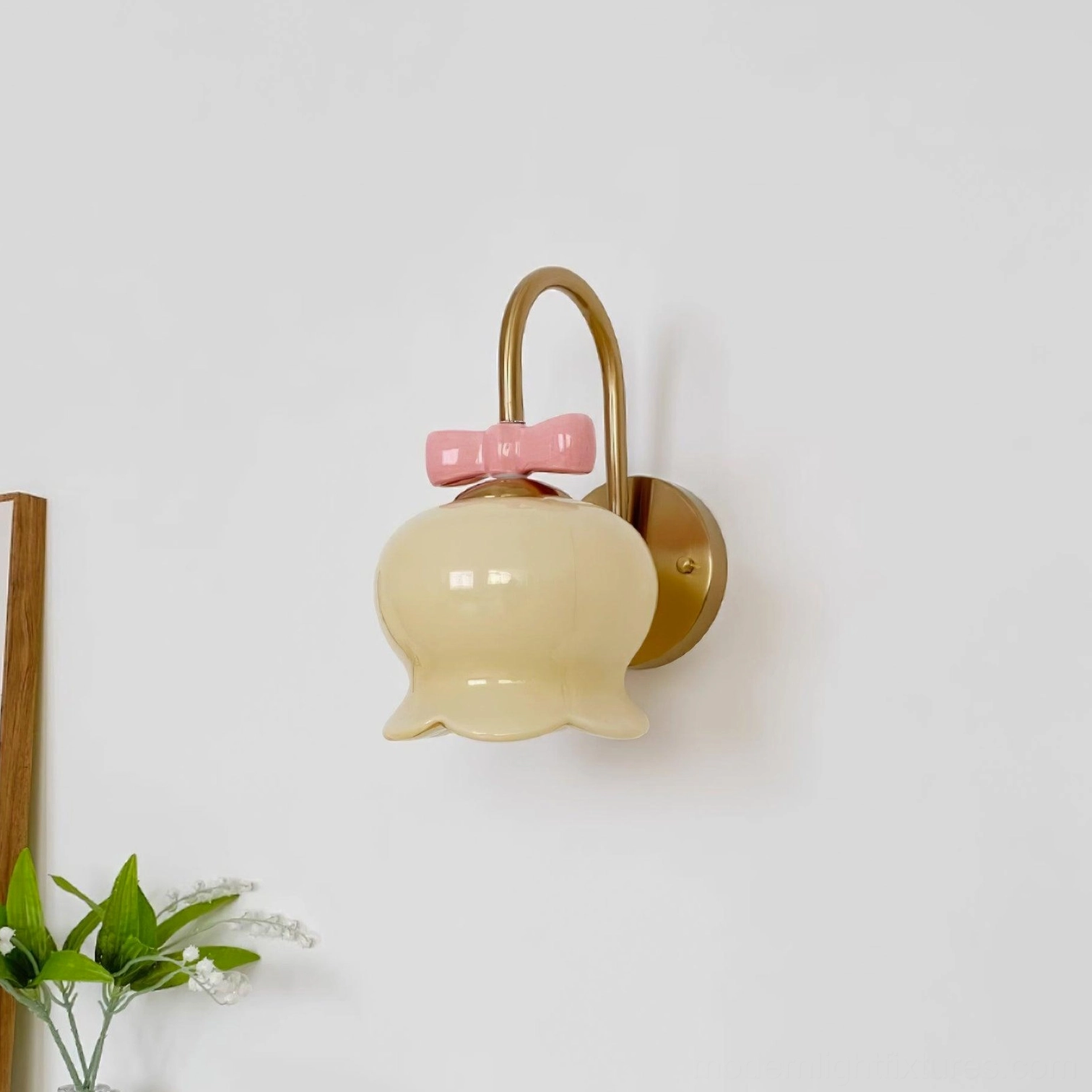Cream yellow ceramic Millie Bells wall sconce with brass metal mounting bracket and pink bow detail, displayed in minimalist home interior with green plants