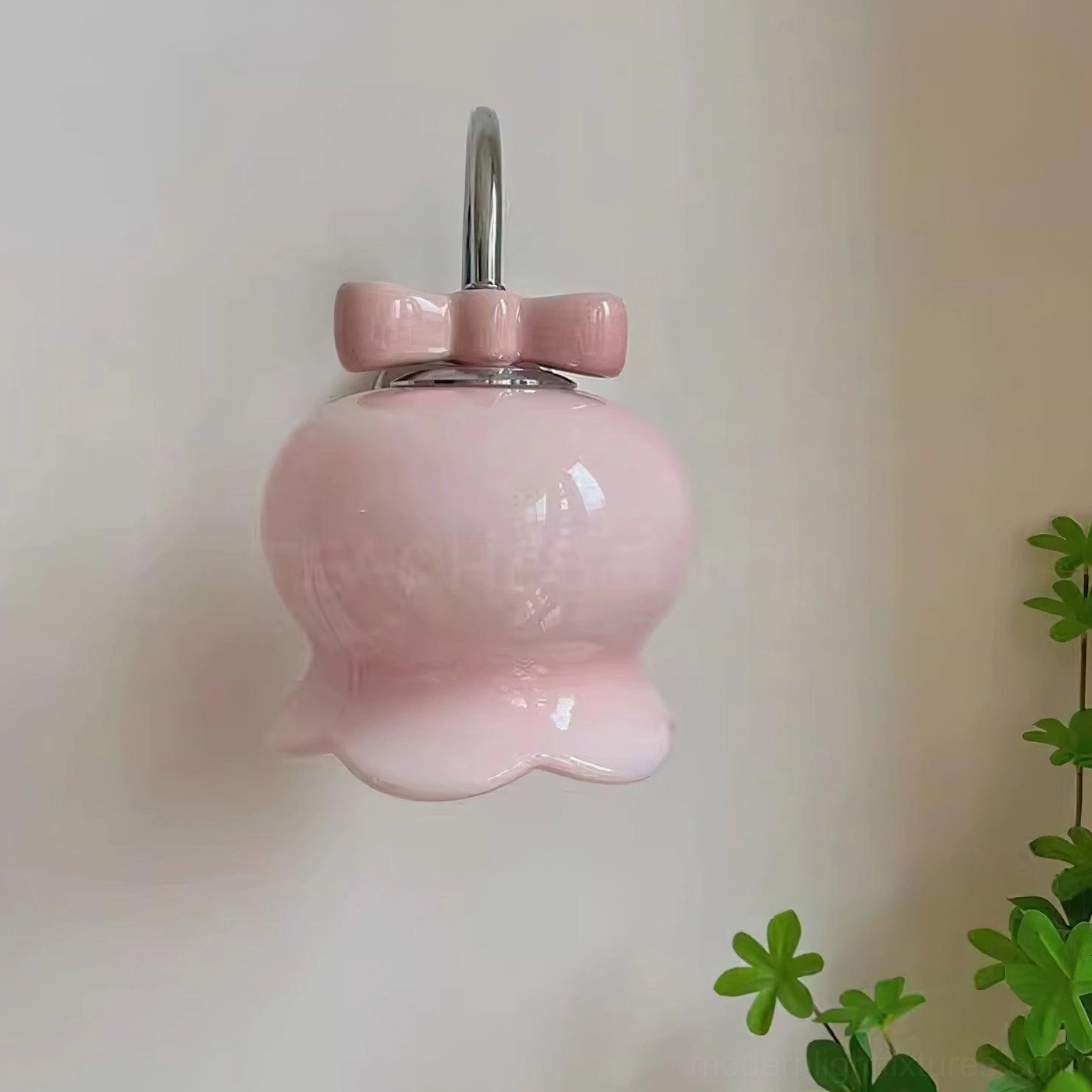 Gradient pink ceramic Millie Bells wall sconce with chrome metal mounting bracket and bow accent, mounted on white wall with green plant decor