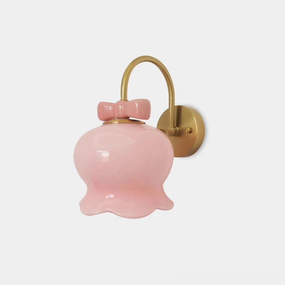 Millie Bells wall sconce with pink ceramic bell shade and brass metal bracket featuring decorative bow accent, front three-quarter studio view