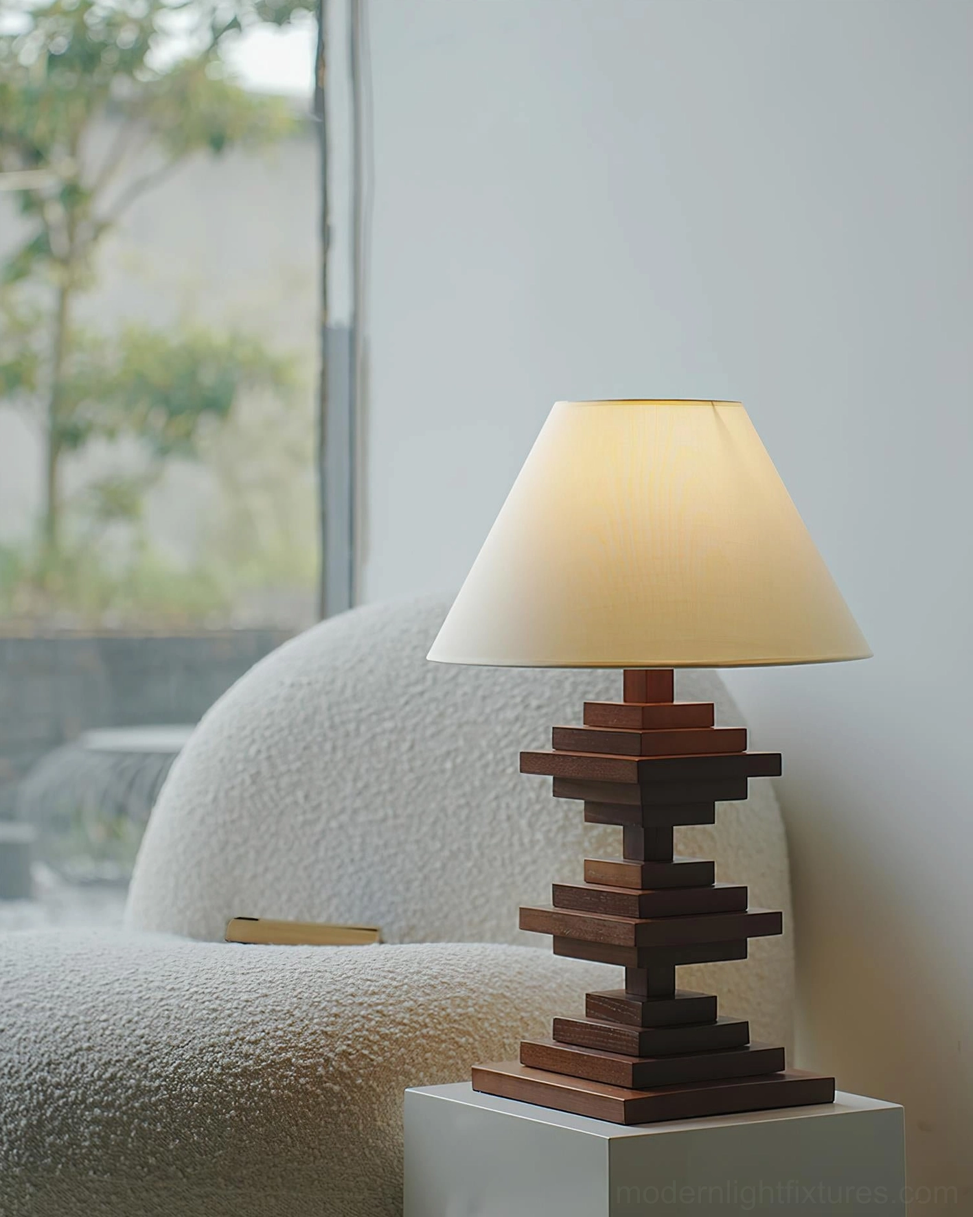 Minimalist wooden tower table lamp near window with grey armchair and white square pedestal showing natural daylight setting
