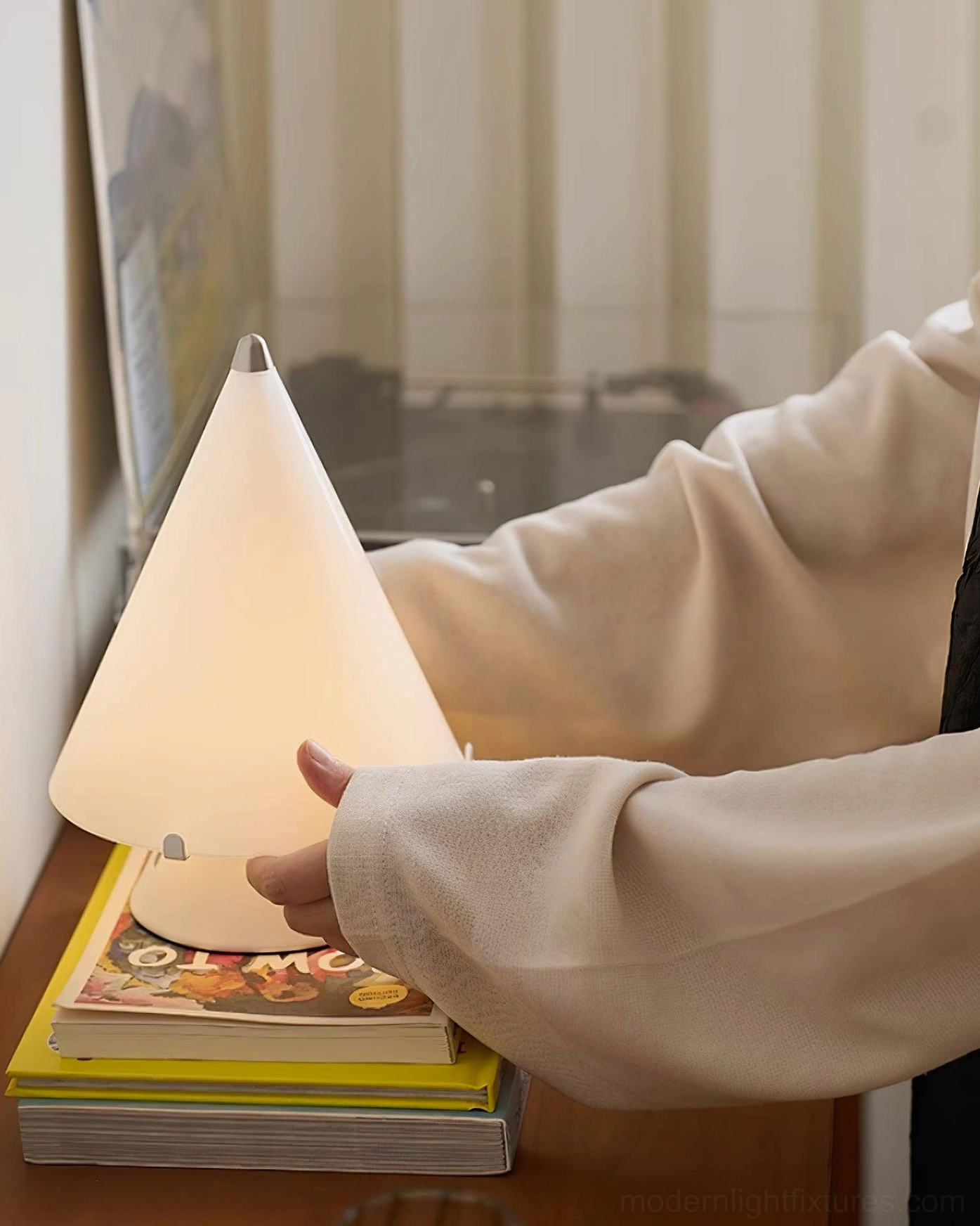 Hand holding white base of Miza minimalist table lamp on book stack with home office background