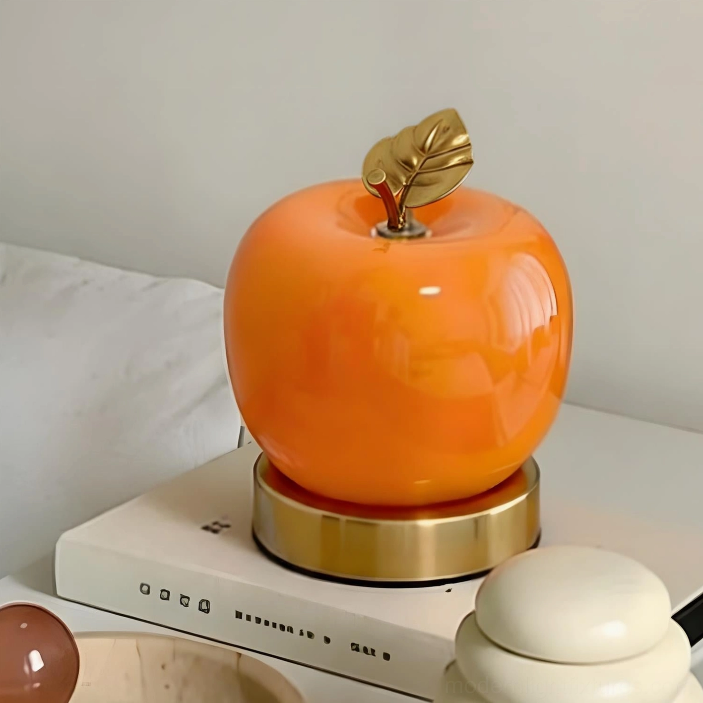Orange apple shaped table lamp placed on bedside table with stacked books, three-quarter view showing sculptural glass body