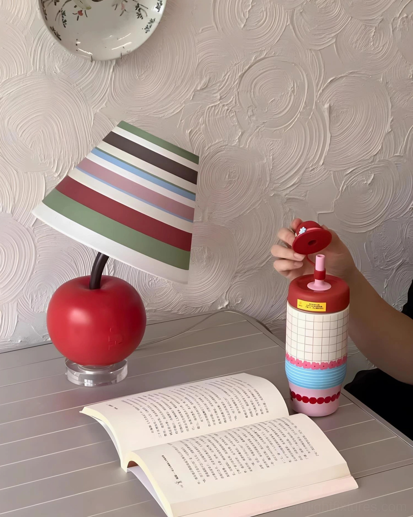 Contemporary apple table lamp with white polka-dot shade on textured wall bedroom desk, front eye-level view with open book