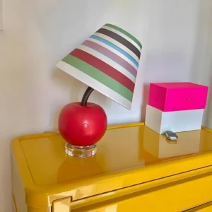 Modern red apple table lamp displayed on yellow wooden dresser with pink-white storage box, front three-quarter perspective